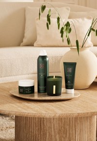 Zestaw zielonych produktów do kąpieli na okrągłej, jasnej marmurowej tacy: świeca, pianka do prysznica, krem do ciała oraz peeling w białych i zielonych opakowaniach.