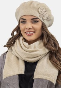 Beige barett s třemi rolovanými látkovými akcenty, spárovaný s odpovídající béžovou šálou, oboje vyrobené z měkkého, strukturovaného materiálu.