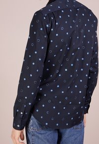 Marinblå skjorta med blå cirkulära mönster, långa ärmar, knäppningar vid ärmsluten och ryggsömn. Tillverkad av ett mjukt, lätt tyg.