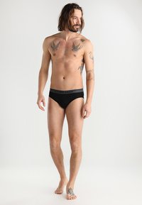 Svarta bomullstrosor med en grå midjeband som har varumärket "Icebreaker" på. Designen är låg midja med platta sömmar för komfort.