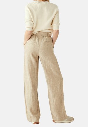 Kobieta w beżowych szerokich lnianych spodniach i kremowym swetrze z prążkowanej dzianiny, z rękami w kieszeniach, stoi tyłem do aparatu.