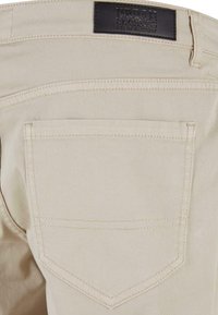 Pantalon en denim beige avec une poche arrière et un patch d’étiquette noire indiquant « Urban Classics Denim Factory » sur la ceinture.