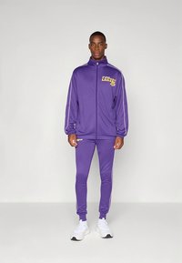 Fato de treino roxo feito de tecido liso com riscas brancas laterais, apresentando um logótipo "Lakers" no casaco e nas calças. Ténis desportivos brancos.