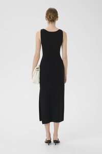 Black sleeveless midi dress with a rounded neckline, side slit, and fitted design. Model holds a cream-colored bag and wears black heeled sandals.
