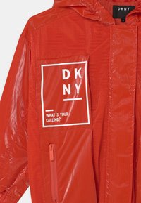 Czerwona kurtka przeciwdeszczowa z panelem z siateczki, kieszenią na zamek i białym nadrukiem tekstowym. Wykonana z błyszczącego, lekkiego materiału. Projekt z kapturem.