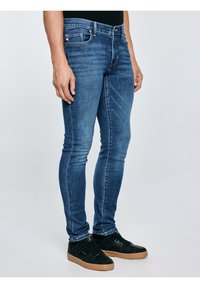 Vīrietis, kas valkā pieguļošas zilas džinsa bikses un melnus sporta apavus ar gumijas zoli, stāv pretvienkāršu gaišu fonu.