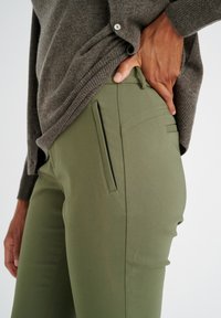 Pantaloni verde oliva aderenti con tasche frontali, realizzati in materiale liscio. La vita è discreta e un maglione grigio a maglia è parzialmente visibile sopra.