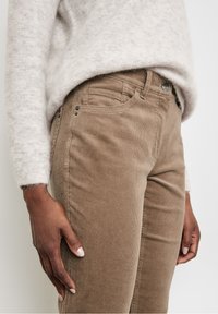 Pantalon en velours côtelé marron avec fines côtes verticales, poches avant et fermeture à bouton métallique, porté avec un pull gris clair pelucheux.