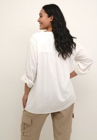 Chemise blanche à manches longues en tissu léger, avec une empiècement au dos et des manches retroussées. Assortie à un pantalon cargo beige.