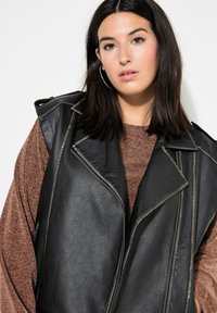 Schwarze Kunstlederweste mit Reverskragen, Reißverschlusselementen und ärmellosem Design, über einem braunen strukturierten Pullover getragen.