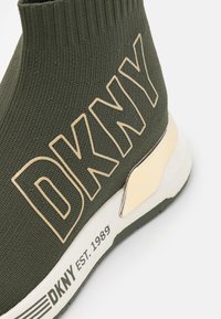 Baskets en maille vert olive avec grands lettrages beiges DKNY, semelle blanche, détail doré au talon et "DKNY EST. 1989" imprimé sur la semelle.
