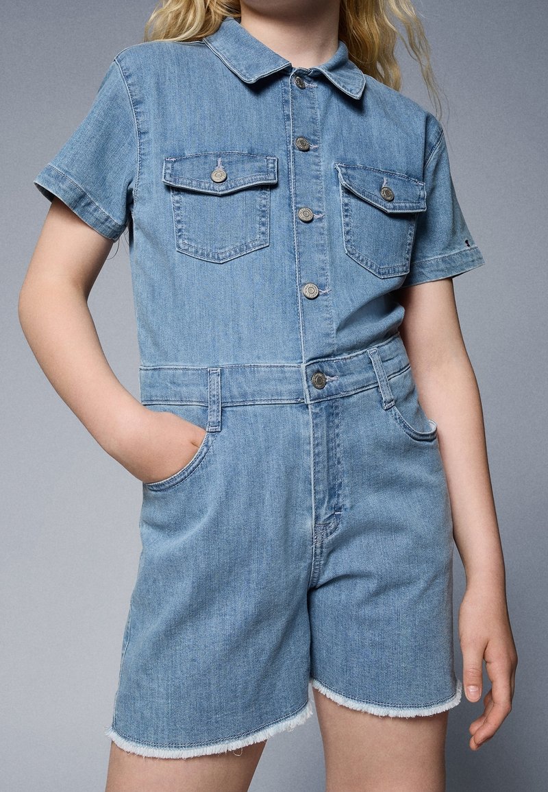 Džínový overal v světle modré barvě, má krátké rukávy, límec, dvě kapsy na hrudi, knoflíky a třepený lem. Materiál působí měkce a odolně.