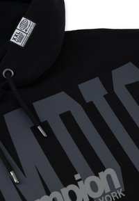 Felpa nera con grande testo grigio e logo. Presenta un cordino con punta metallica e un'etichetta in tessuto con informazioni sulla taglia. Texture morbida.