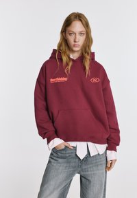 Bordó kapucnis pulóver elülső zsebbel és bordázott mandzsettával, rózsaszín "Overthinking" felirattal és grafikus logóval, világos csíkos ing fölött viselve.