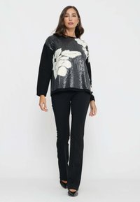 Maglione nero con paillettes e design floreale bianco, abbinato a pantaloni neri e scarpe nere a punta, modello in piedi davanti a uno sfondo neutro.