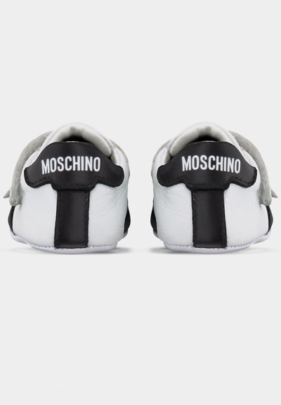Λευκά και μαύρα παιδικά αθλητικά παπούτσια, ορατά από πίσω, το καθένα με το "MOSCHINO" τυπωμένο με λευκά γράμματα στη μαύρη ετικέτα της φτέρνας.