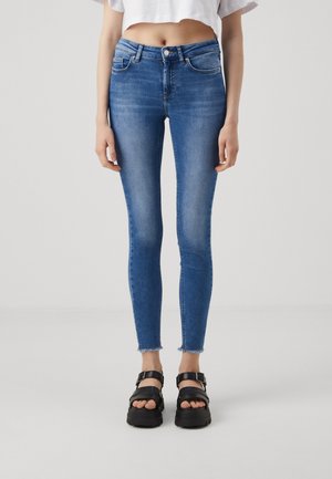 Vrouw draagt mid-rise blauwe skinny jeans met onafgewerkte zoom en zwarte platformsandalen, staand tegen een effen lichte achtergrond.