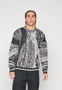 Strickpullover mit einem strukturierten Schwarz-Weiß-Muster, das unterschiedliche Kabel- und geometrische Designs aufweist. Rippbündchen und Saum, Etikett am unteren Rand.