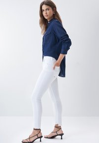 Camisa de mezclilla abotonada en azul oscuro, combinada con jeans skinny blancos. El modelo lleva sandalias de tacón alto negras con tiras. Diseño simple y minimalista.