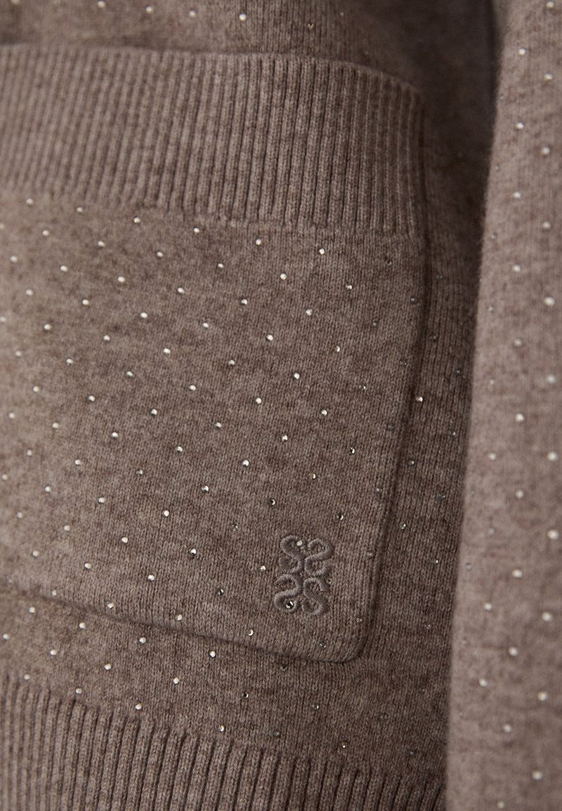 Brauner Strickpullover mit strukturierter Oberfläche, verziert mit kleinen silbernen Nieten, gerippten Bündchen und einem Logo-Patch auf der Tasche.