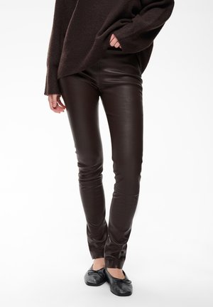 Person trägt dunkelbraune Lederhose, schwarze Ballerinas und einen lockeren dunkelbraunen Pullover mit einer Hand in der Tasche.