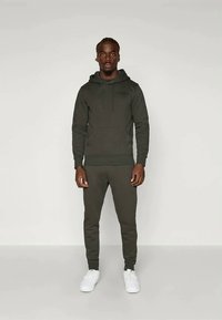 Σκούρο πράσινο φούτερ με καγκουρό τσέπη, συνδυασμένο με αντίστοιχες φόρμες και λευκά αθλητικά παπούτσια. Διαθέτει κουκούλα από κορδόνι και ραμμένα μανίκια.