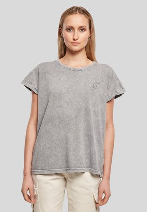 Femme portant un t-shirt ample gris à manches courtes avec un petit motif cerise sur la poitrine et un pantalon cargo beige clair, debout devant un fond uni.