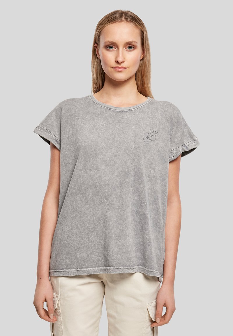 Femme portant un t-shirt ample gris à manches courtes avec un petit motif cerise sur la poitrine et un pantalon cargo beige clair, debout devant un fond uni.