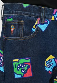 Jeans din denim cu petice grafice colorate, inclusiv fețe în nuanțe de albastru, verde, roz și galben, cu cusături aurii și un finisaj texturat.