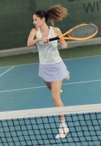 Jugadora de tenis femenina con una camiseta sin mangas blanca y una falda morada clara, golpeando una raqueta naranja en una cancha azul, con una red en primer plano.