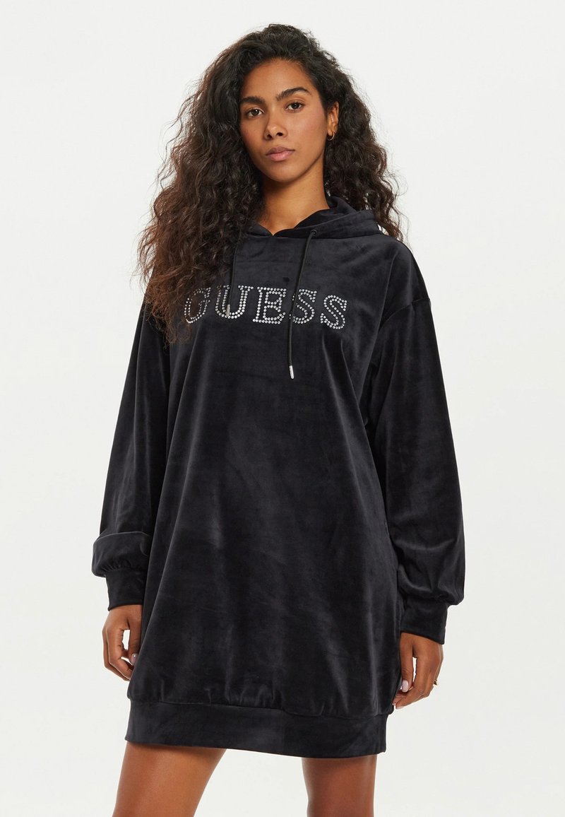 Abito felpa nero in velour con logo "GUESS" in strass sul davanti, maniche lunghe e una vestibilità rilassata con orlo appena sopra il ginocchio.