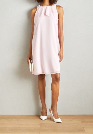 Femme portant une robe rose pâle sans manches longueur genou, tenant une pochette dans la main droite et portant des chaussures blanches à talons hauts.