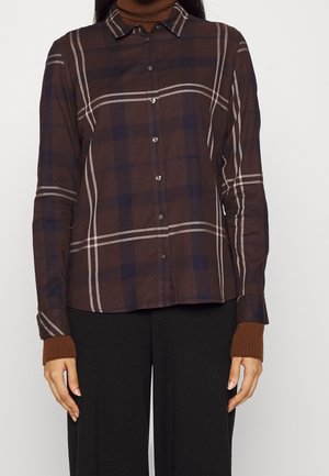 Kobieta w brązowo-granatowej koszuli w kratę zapinanej na guziki, nałożonej na brązowy sweter z golfem oraz czarne spodnie, stoi na jednolitym tle.