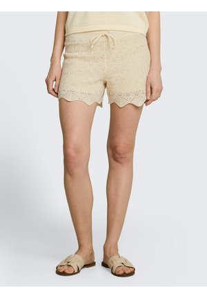 Kobieta w beżowych dzianinowych szortach z falowanym obszyciem i sznurkiem w pasie, w połączeniu z beżowymi sandałami typu slip-on.