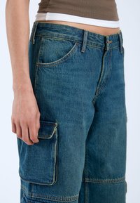Pantalones cargo de mezclilla en azul medio con un corte relajado, que presentan grandes bolsillos laterales y detalles de costura visibles.