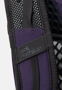 Sac à dos violet avec un panneau en maille noire, des sangles grises et une étiquette de marque. Tissu texturé et construction durable visibles en détail.