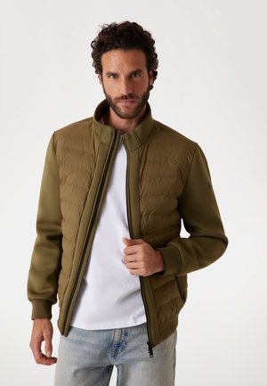 Chaqueta verde oliva con frontales acolchados, mangas suaves, cuello alto y cierre de cremallera. Llevada sobre una camisa blanca con denim claro.