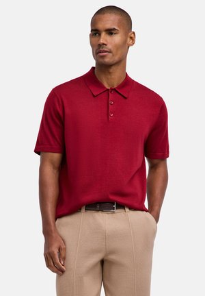 Uomo che indossa una polo a maniche corte di colore rosso scuro infilata in pantaloni beige con una cintura nera, in piedi con una mano in tasca.
