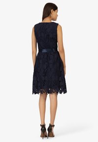 Robe en dentelle marine avec un corsage ajusté, un ourlet festonné et une ceinture en satin. Portée avec des chaussures à talons noirs, mettant en valeur des motifs floraux complexes.