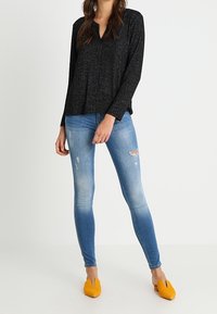 Žena v černém dlouhém rukávu, světlých ošoupaných skinny džínách a žlutých špičatých nazouvácích stojí na jednobarevném pozadí.