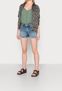 Chaqueta bomber floral con ribete negro, camisa verde de botones, pantalones cortos de mezclilla desgastados y sandalias marrones con correas. Atuendo casual de verano.
