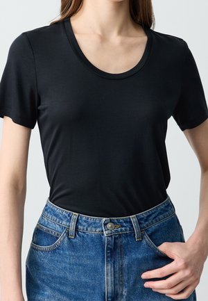 Woman wearing a plain black short-sleeve round-neck t-shirt tucked into high-waisted blue denim jeans with one hand in pocket.