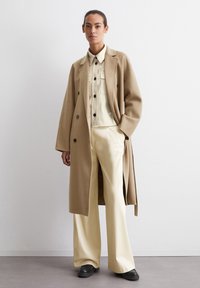 Cappotto in lana beige chiaro, doppiopetto, indossato su una camicia crema e pantaloni a gamba larga. Bottoni neri, dettagli di design minimi, texture liscia.