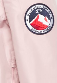 Manga de chaqueta rosa claro con un parche bordado circular que representa montañas en rojo y blanco, con el texto "Cortina D'Ampezzo."