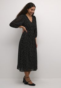 Vestido negro de lunares con escote en V, material semitransparente, mangas globo y cintura con lazo, que llega a la mitad de la pantorrilla, combinado con tacones negros.