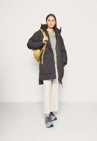 Abrigo negro acolchado con capucha y múltiples bolsillos frontales, camiseta gris clara, pantalones anchos beige y zapatillas azules con detalles negros.