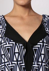 Wielokolorowa bluzka z geometrycznym wzorem w czerni, bieli i niebieskim, z dekoltem w kształcie V oraz czarnymi akcentami przy kołnierzu. Gładka faktura.