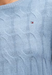 Šviesiai mėlynas megztinis su iškilusiu pynimo raštu ir audiniu apykakle. Yra mažas logotipas ant krūtinės. Minkšta tekstūra.