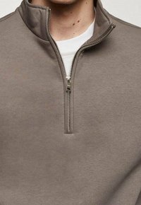 Pullover en tissu gris atténué avec un col zippé à quart. Présente une texture lisse et une fermeture éclair argentée, superposé sur une chemise blanche.