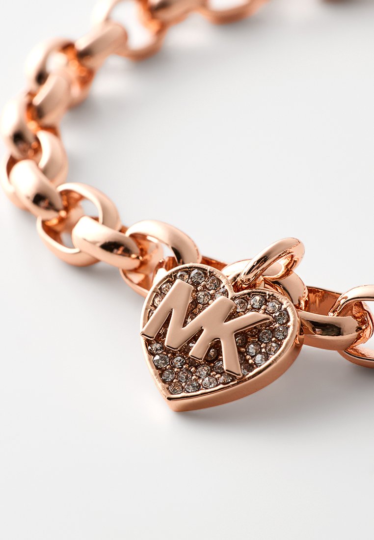 Armband aus roségoldfarbenem Material mit einem Herzanhänger, der "MK" in poliertem Metall zeigt und mit klaren Rhinsteinen verziert ist.
